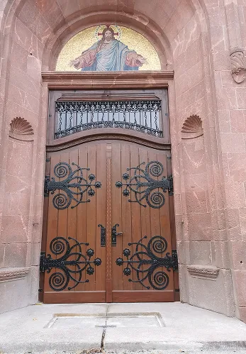 Große Holztür mit dekorativen schwarzen Metallbeschlägen und Jesus-Mosaik darüber in einem Backsteinhaus.