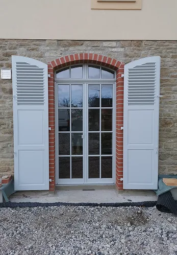 Tür mit geöffnetem weißem Fensterladen und Gitter, umrahmt von einem roten Ziegelbogen.